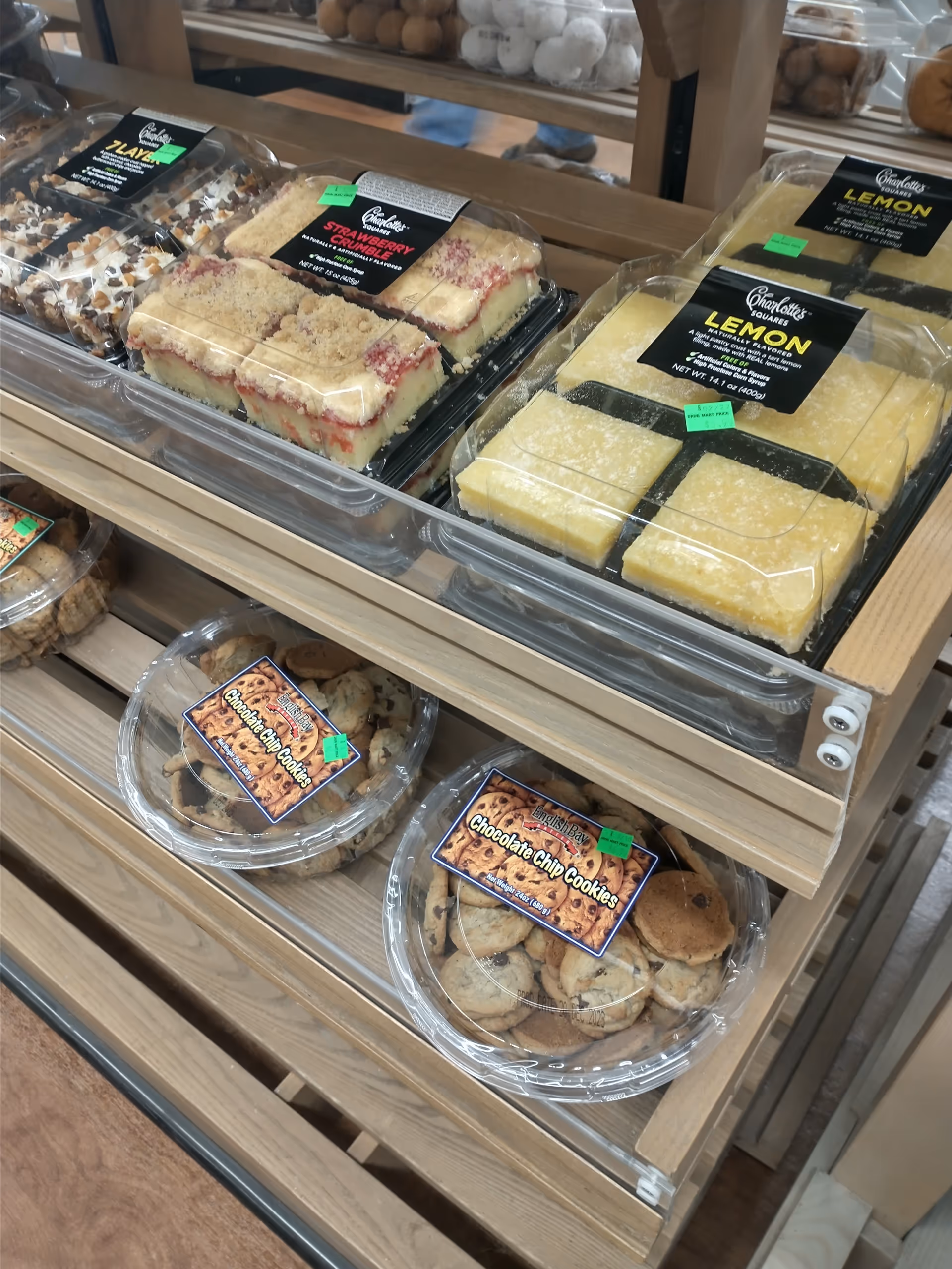 Assorted cakes and cookies in clear trays on wooden shelves inside a shop.