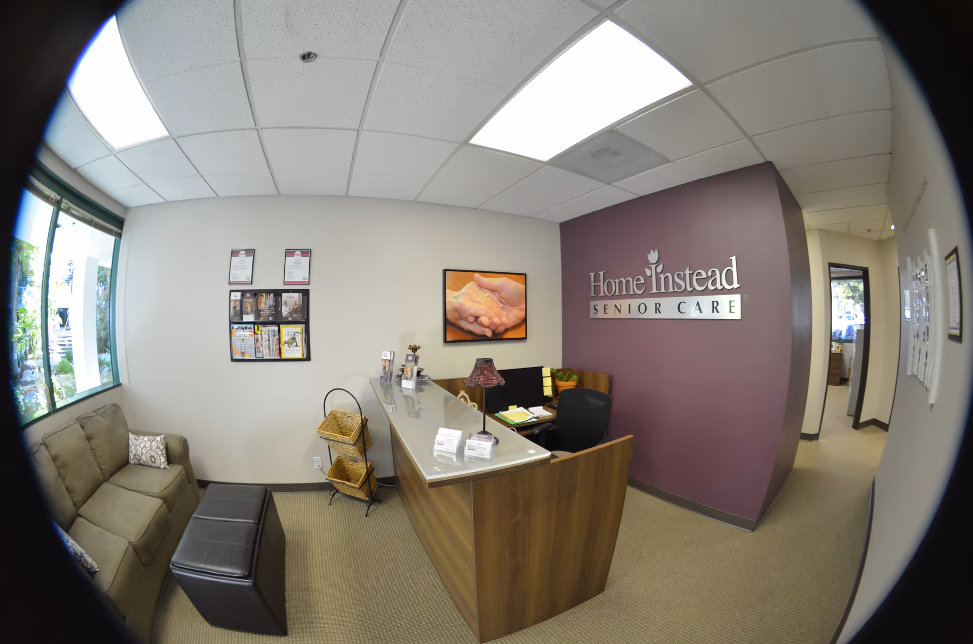 Reception area with a wooden desk, beige seating, and a purple Home Instead wall logo.