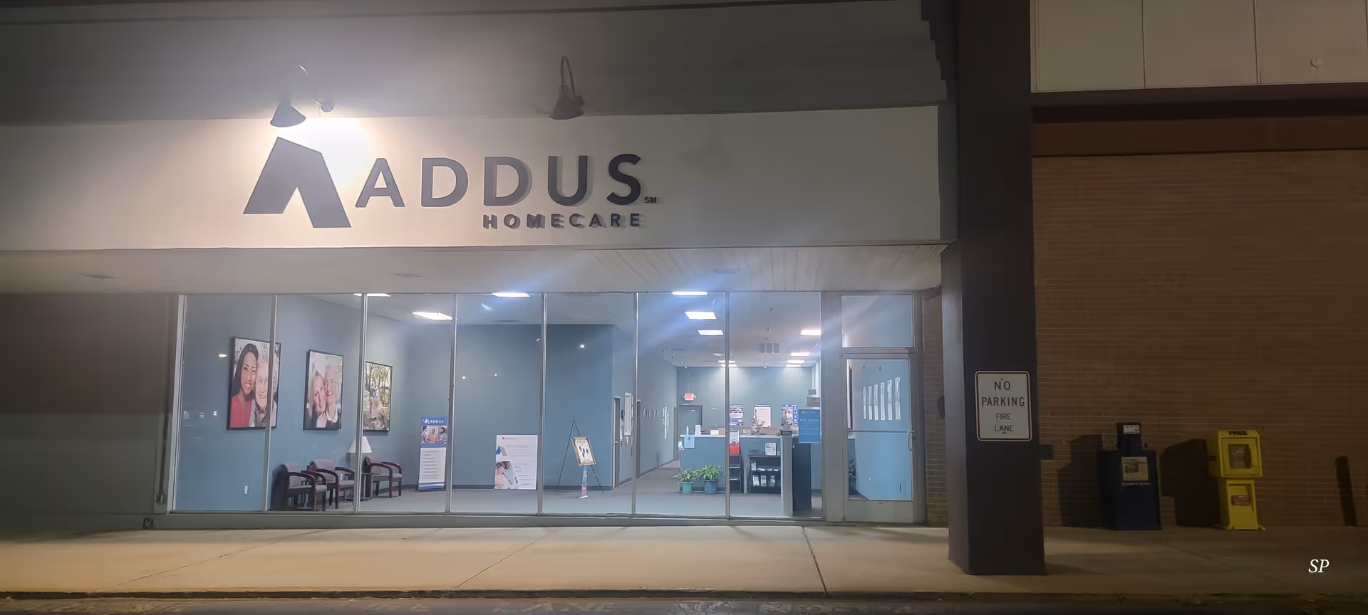 Nighttime view of Addus HomeCare storefront with glass windows and lit interior.