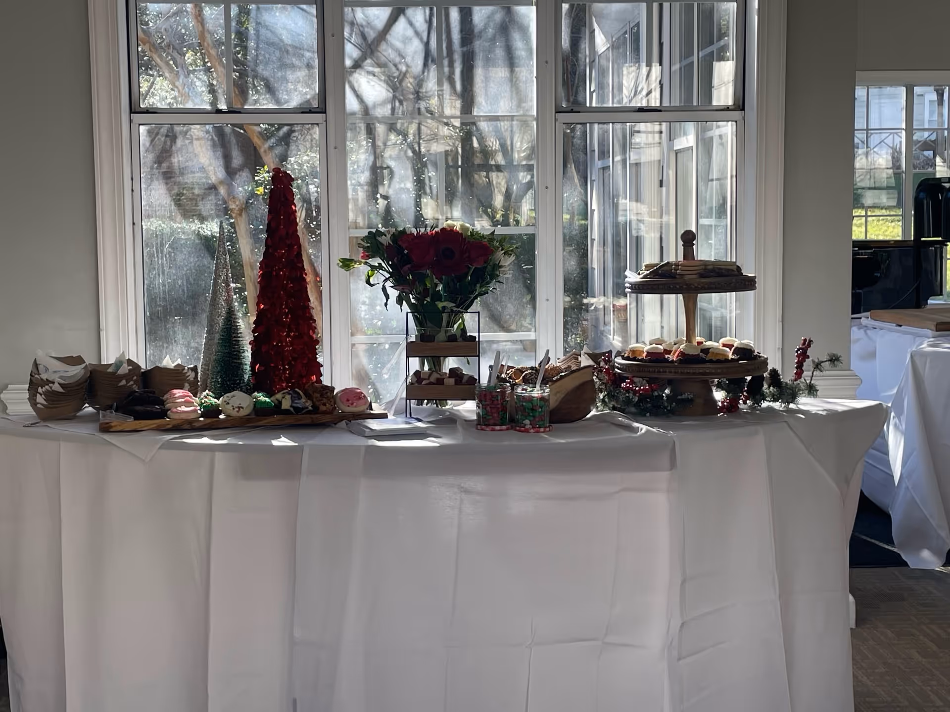 A long pastry table with cupcakes, desserts, and flower arrangements in a bright, windowed room.