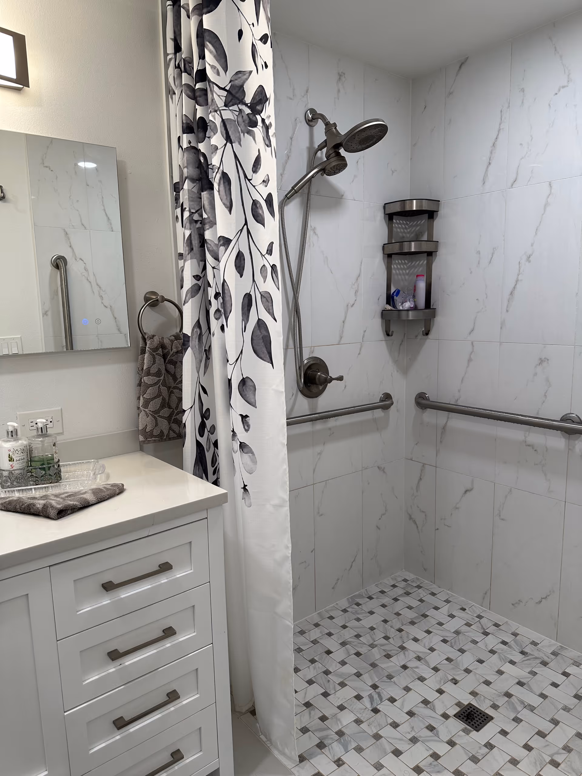 A handicap-accessible bathroom with marble walls, grab bars, a patterned shower curtain, and a white vanity.