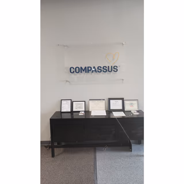 An interior wall features a Compassus sign above a black console table with certificates.