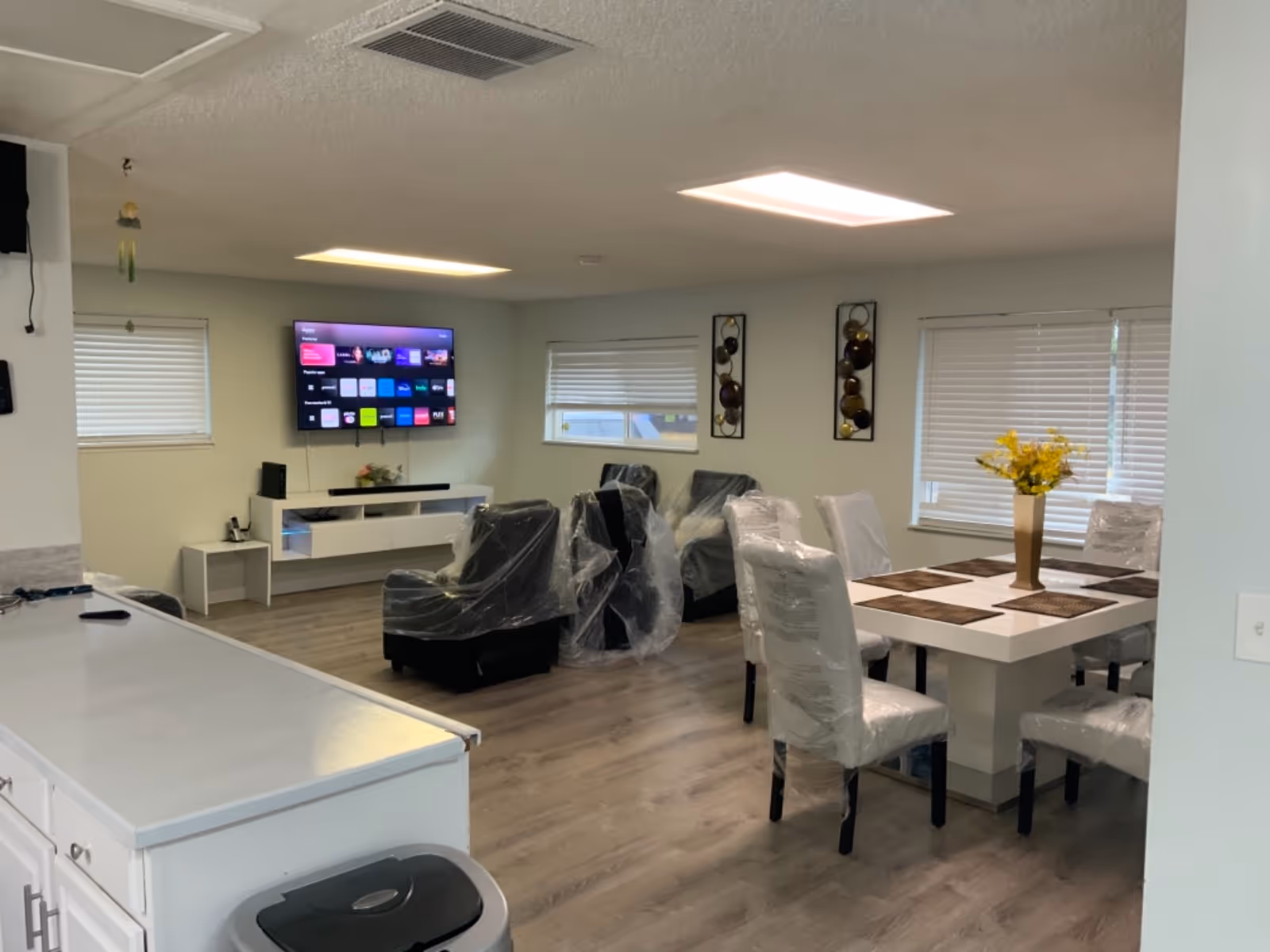 Senior living facility common area with TV, wrapped chairs, dining table, and counter.