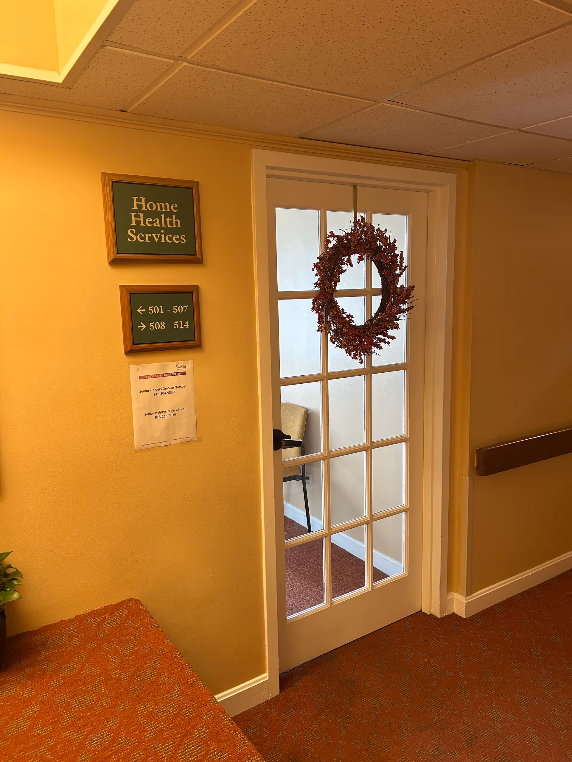 Hallway with a glass-paned door, wreath, and wall signs in a senior living facility.