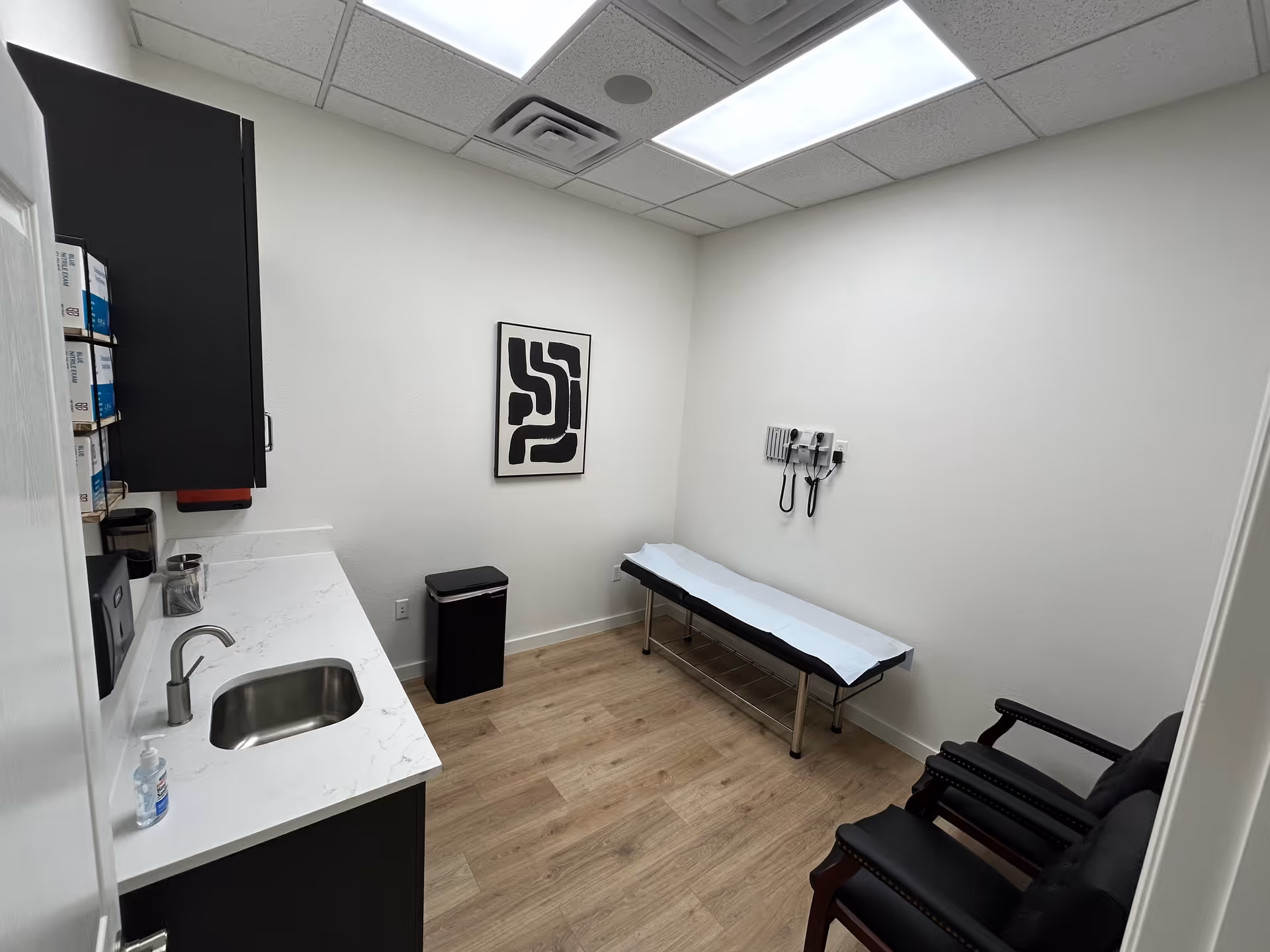 A small medical examination room with sink, cabinets, exam table, stethoscope, and black chairs.