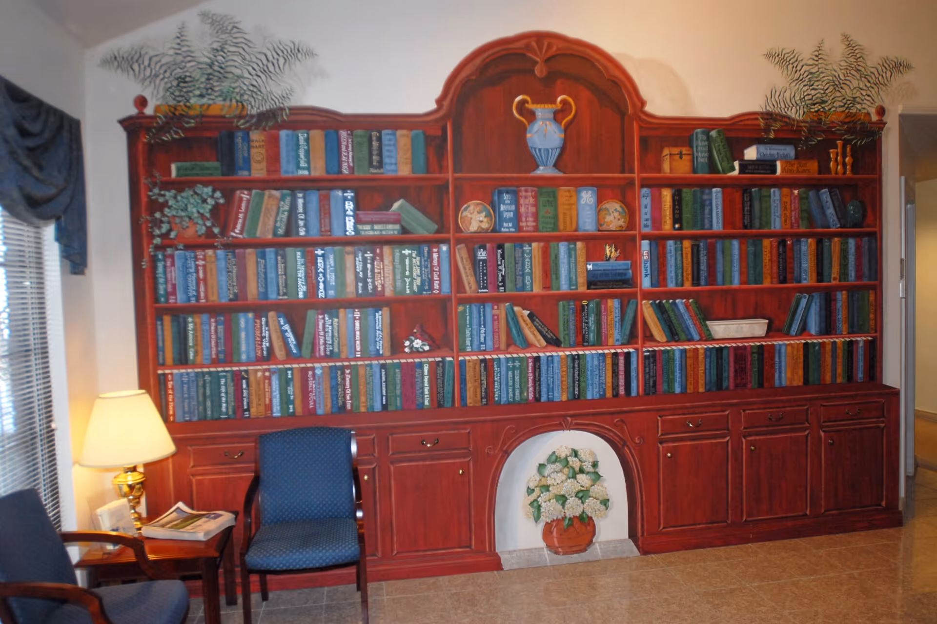 Red wooden floor-to-ceiling bookshelf filled with books, seating area, lamp, decorative fireplace alcove