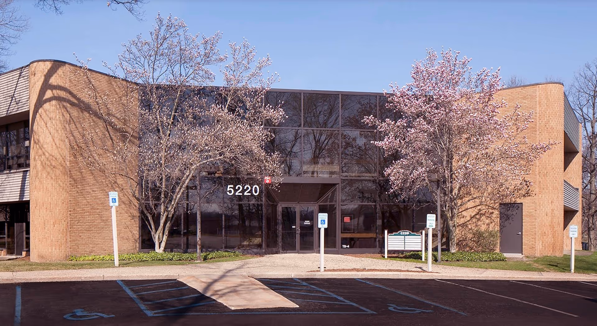 Brick Compassus facility with large glass entry, blooming trees, and a parking lot.