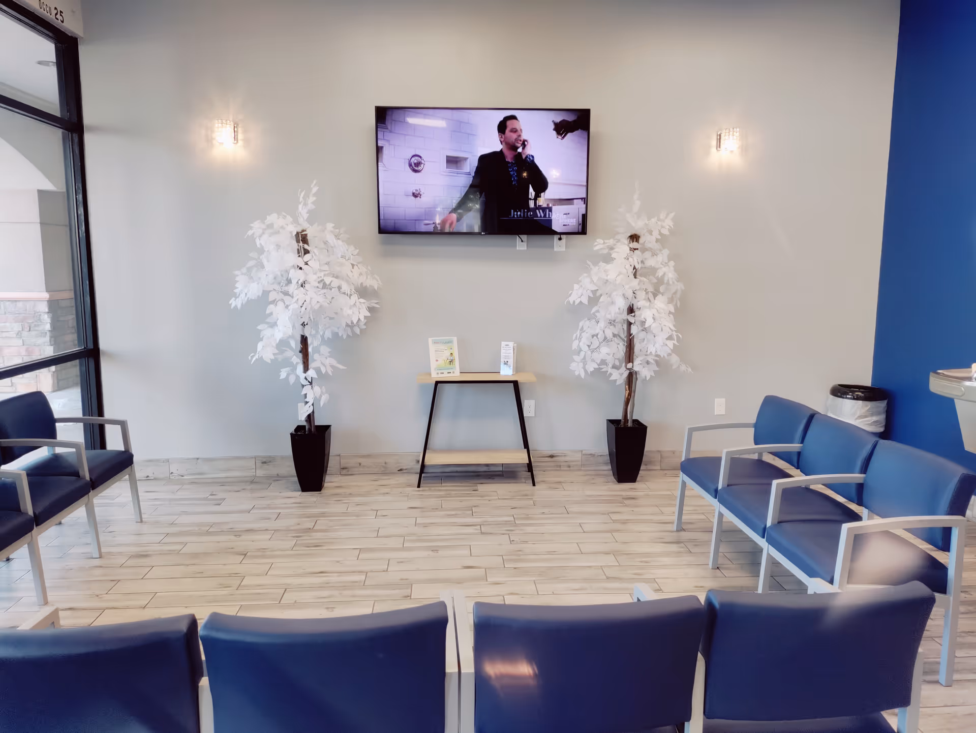 A modern senior living lobby with blue chairs, a wall TV, and decorative trees.