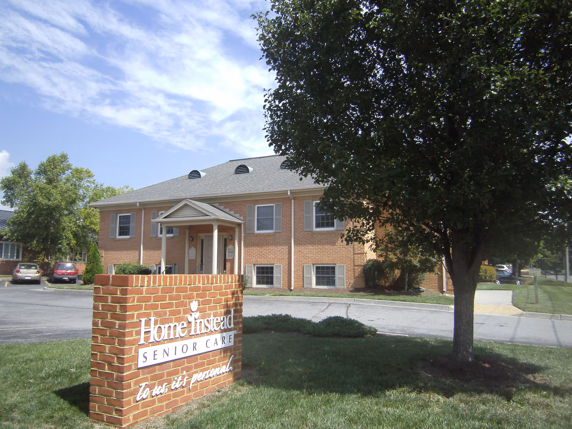 Brick Home Instead Senior Care building with front entrance sign, parking lot, and a large tree.