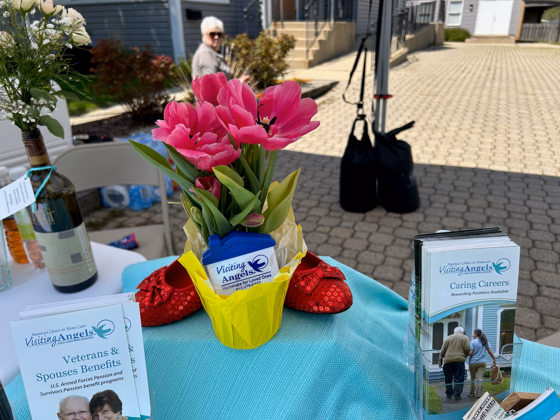 Outdoor promotional table with Visiting Angels brochures, pink flowers, and a blurred passerby.