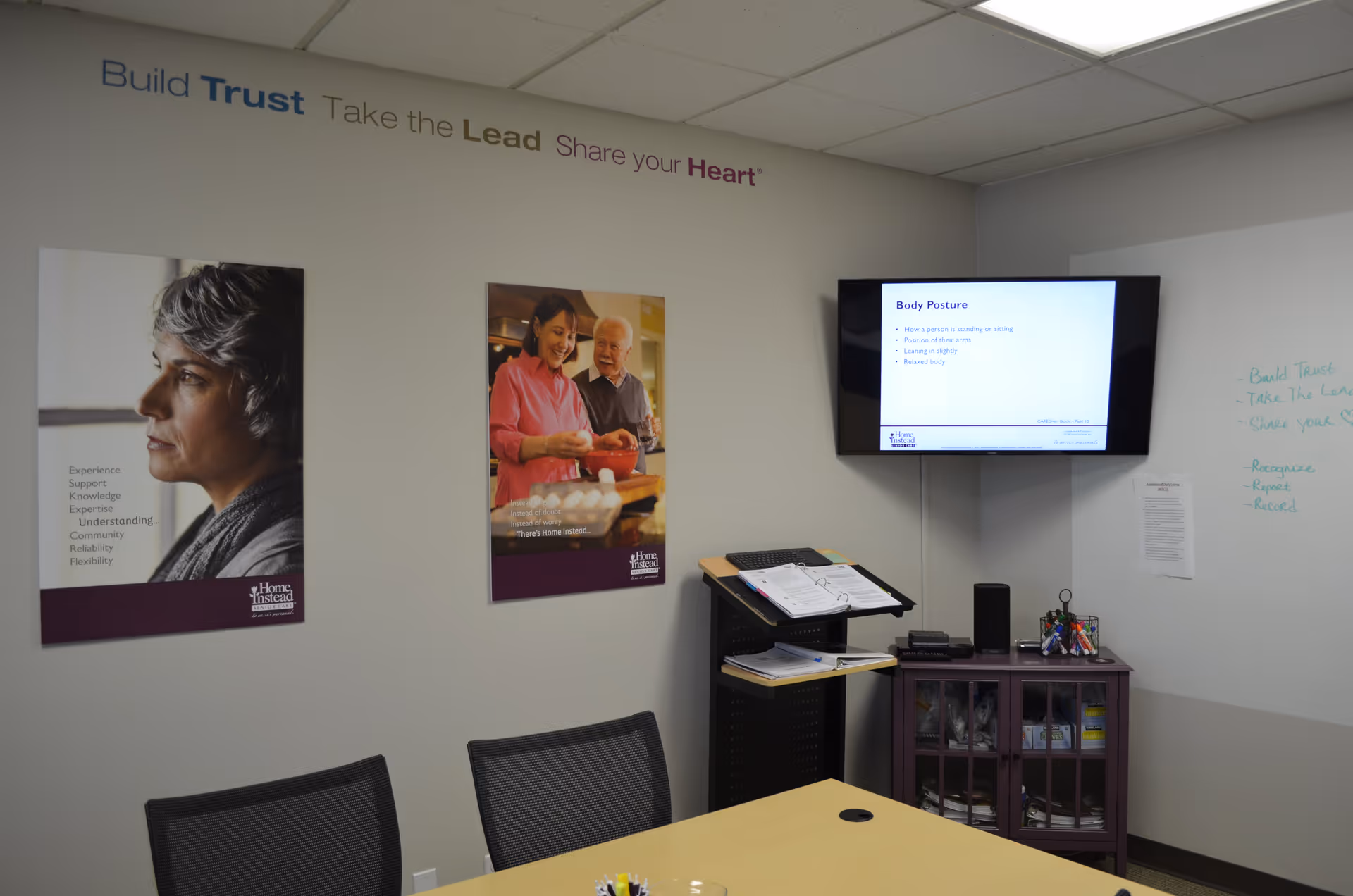 A meeting room in a Home Instead facility with posters, a wall monitor, and a table.