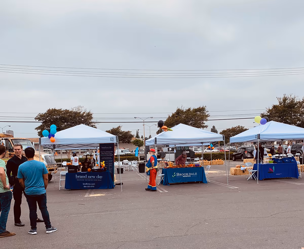 Outdoor community fair with white tents, balloons, tables, and people mingling in a parking lot.