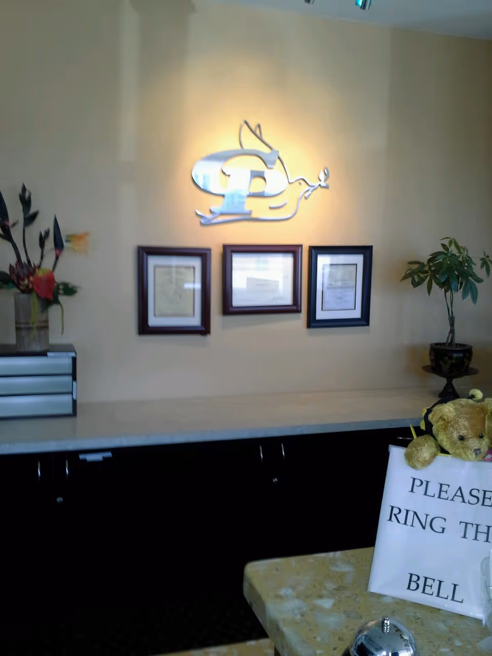 Reception area with logo, framed certificates, plants, and a bell sign on the counter.