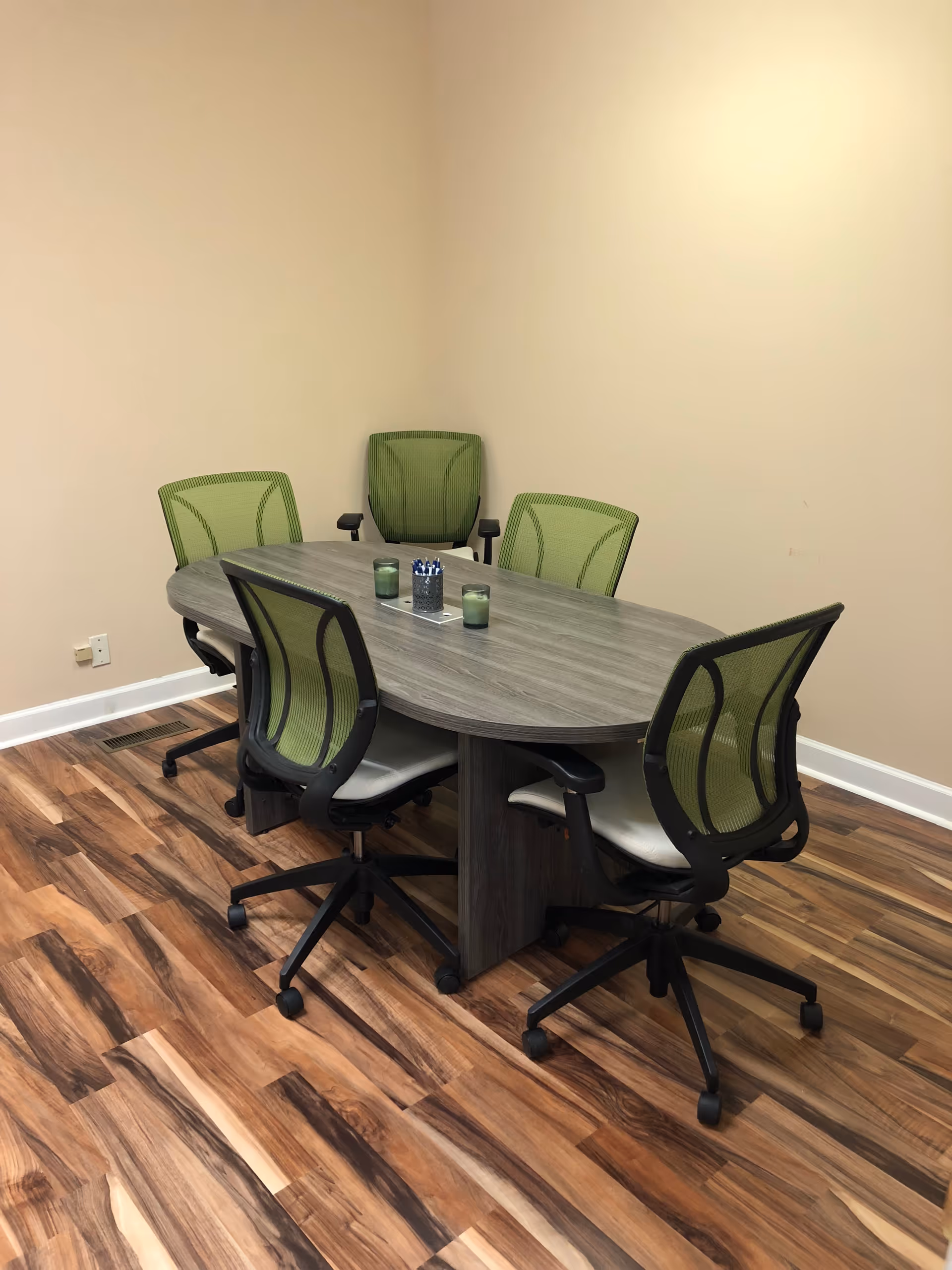 An interior meeting room with a gray oval table on wood floor and green office chairs.