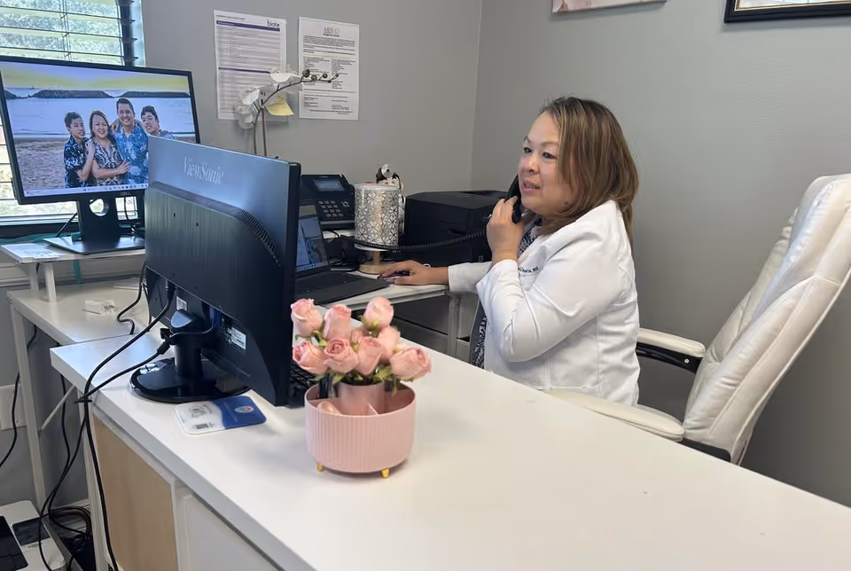 Healthcare professional in white coat at a desk with computer in clinic office.