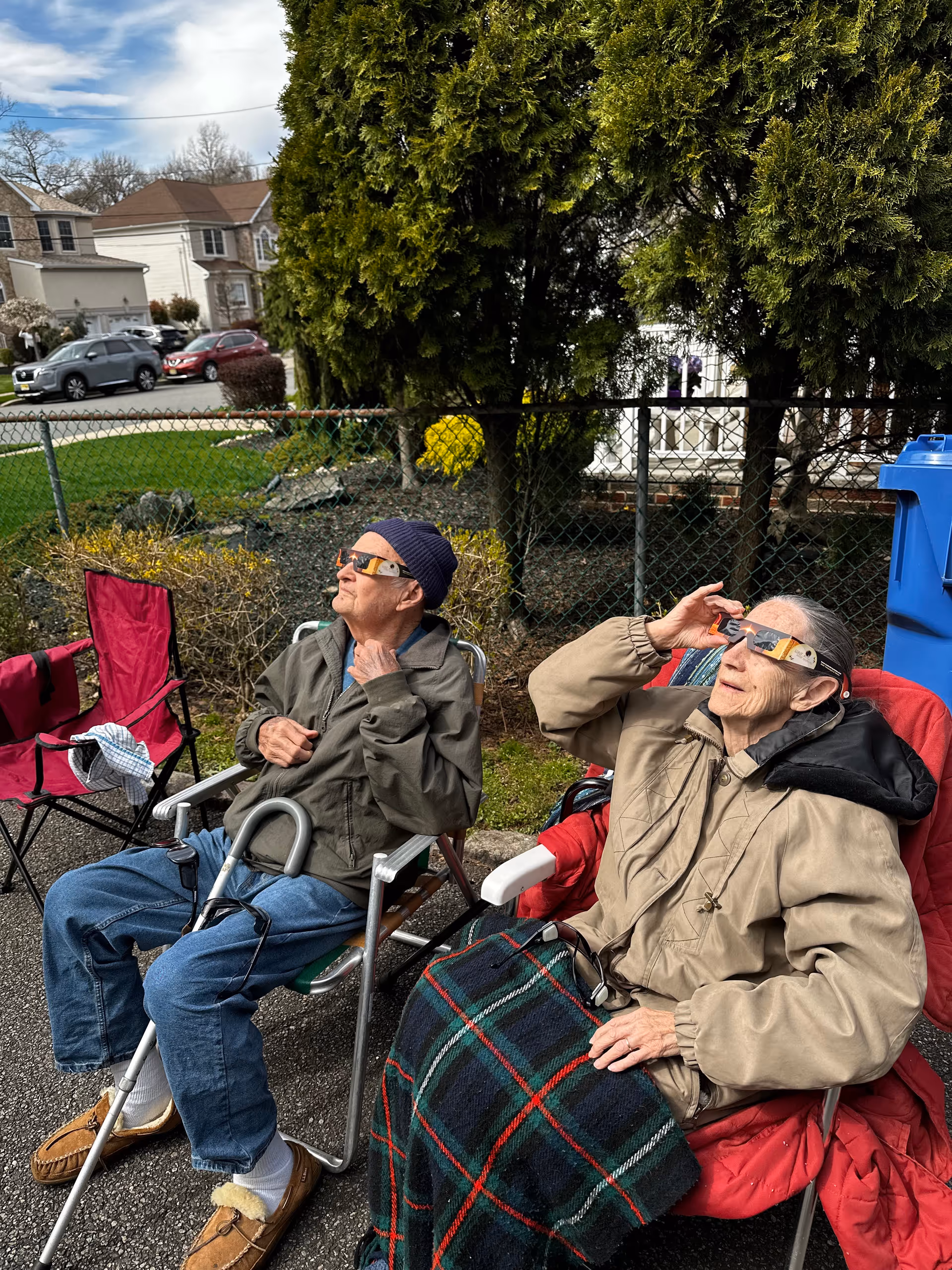 Two elderly residents sit in wheelchairs outside, wearing sunglasses near a chain-link fence.