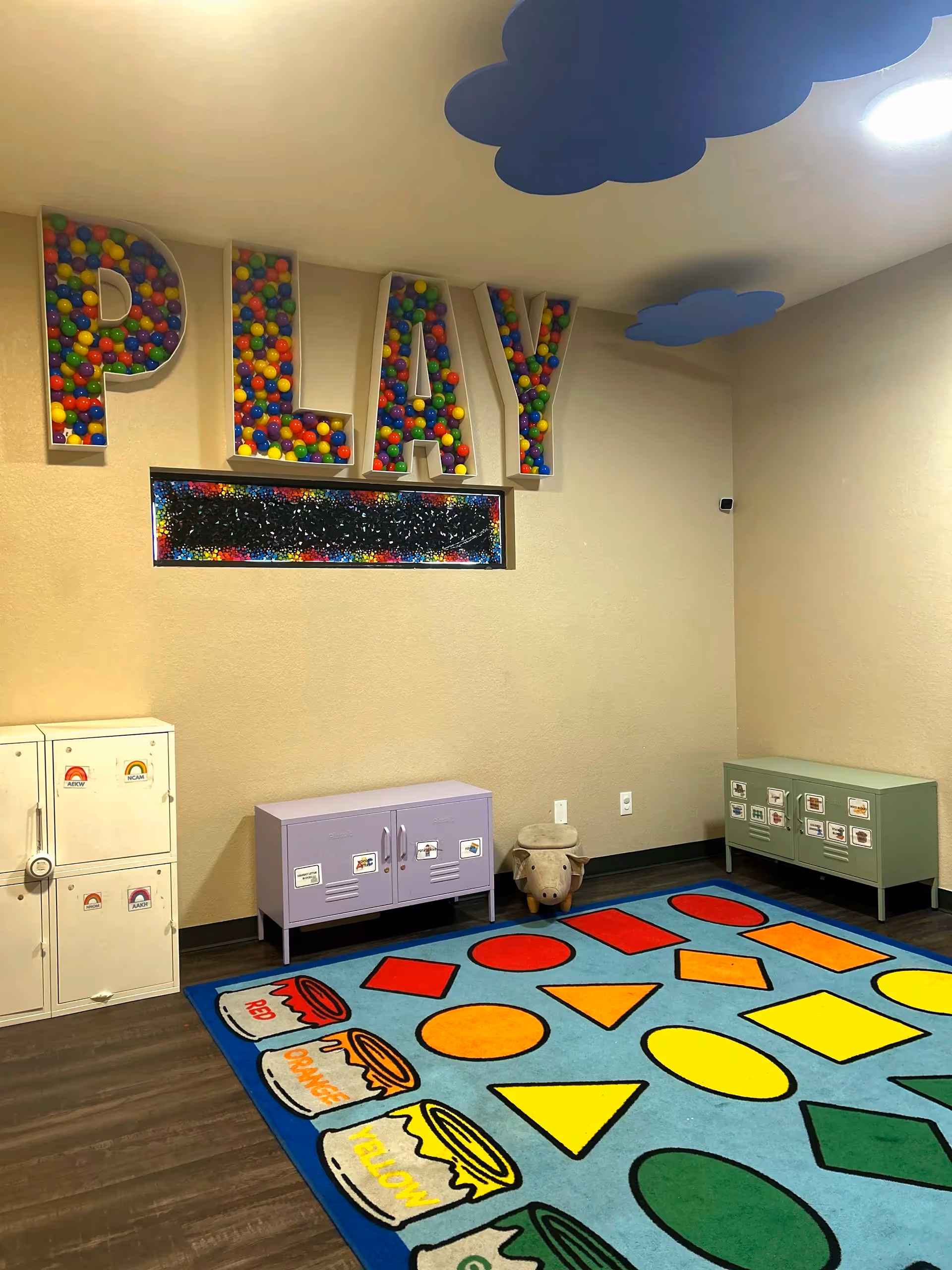 Bright playroom with large rainbow 'PLAY' letters, colorful rug, and cubby storage.