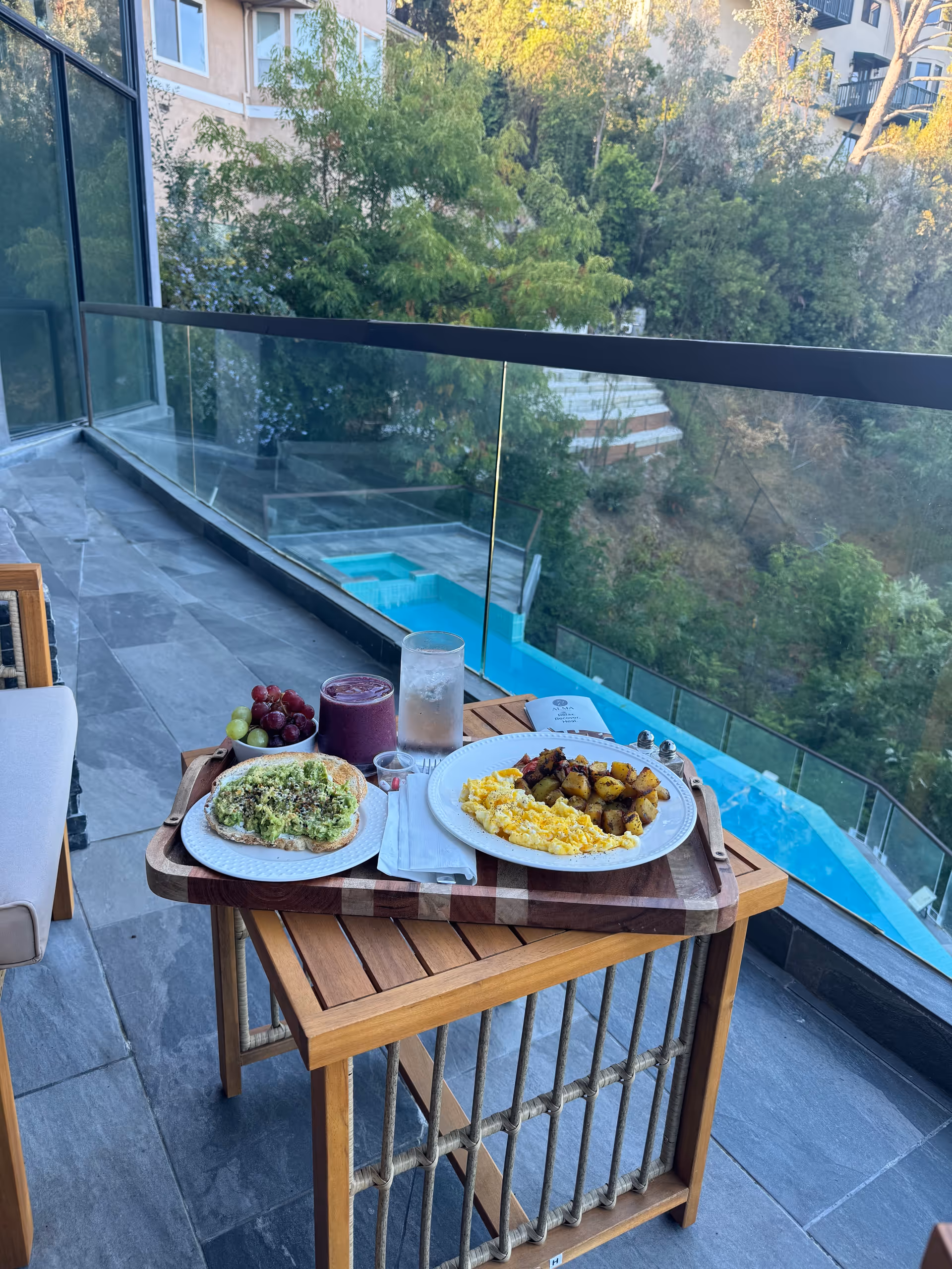 Outdoor balcony meal setup with two plates, fruit, drinks, and glass railing overlooking pool.