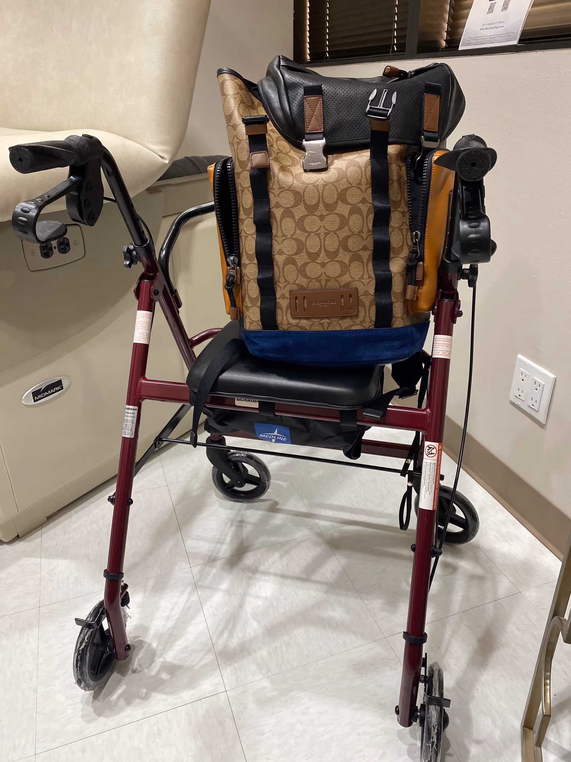 A burgundy wheeled walker with a tan backpack sits in a clinical interior.