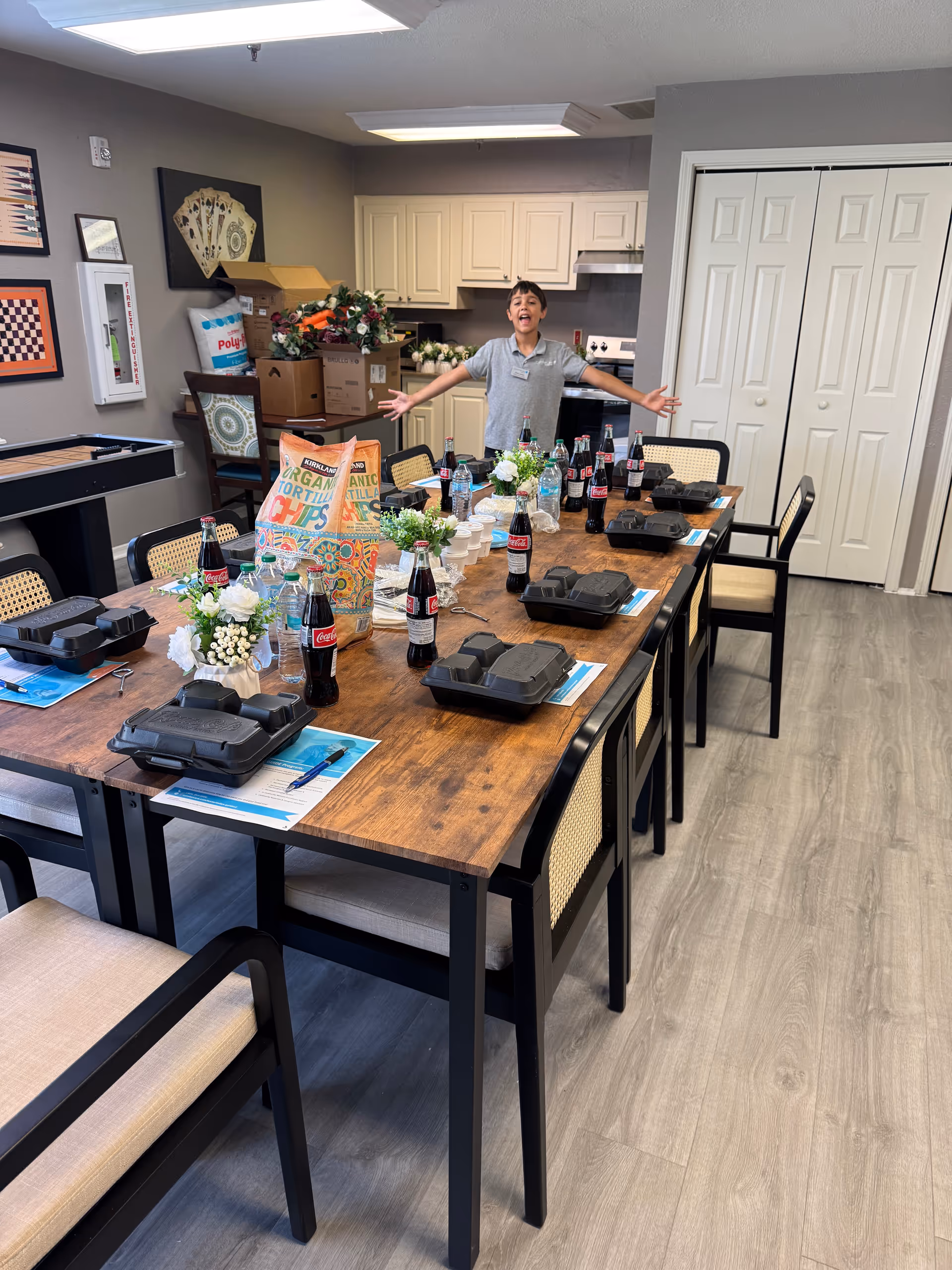 Staff member stands with arms wide beside a long dining table set with takeout boxes.
