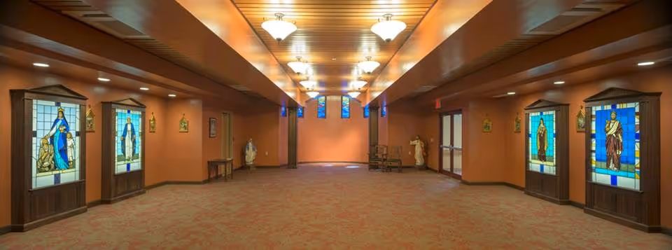 Wide interior hallway in a hospice facility with orange walls, carpet, ceiling lights, and stained glass.
