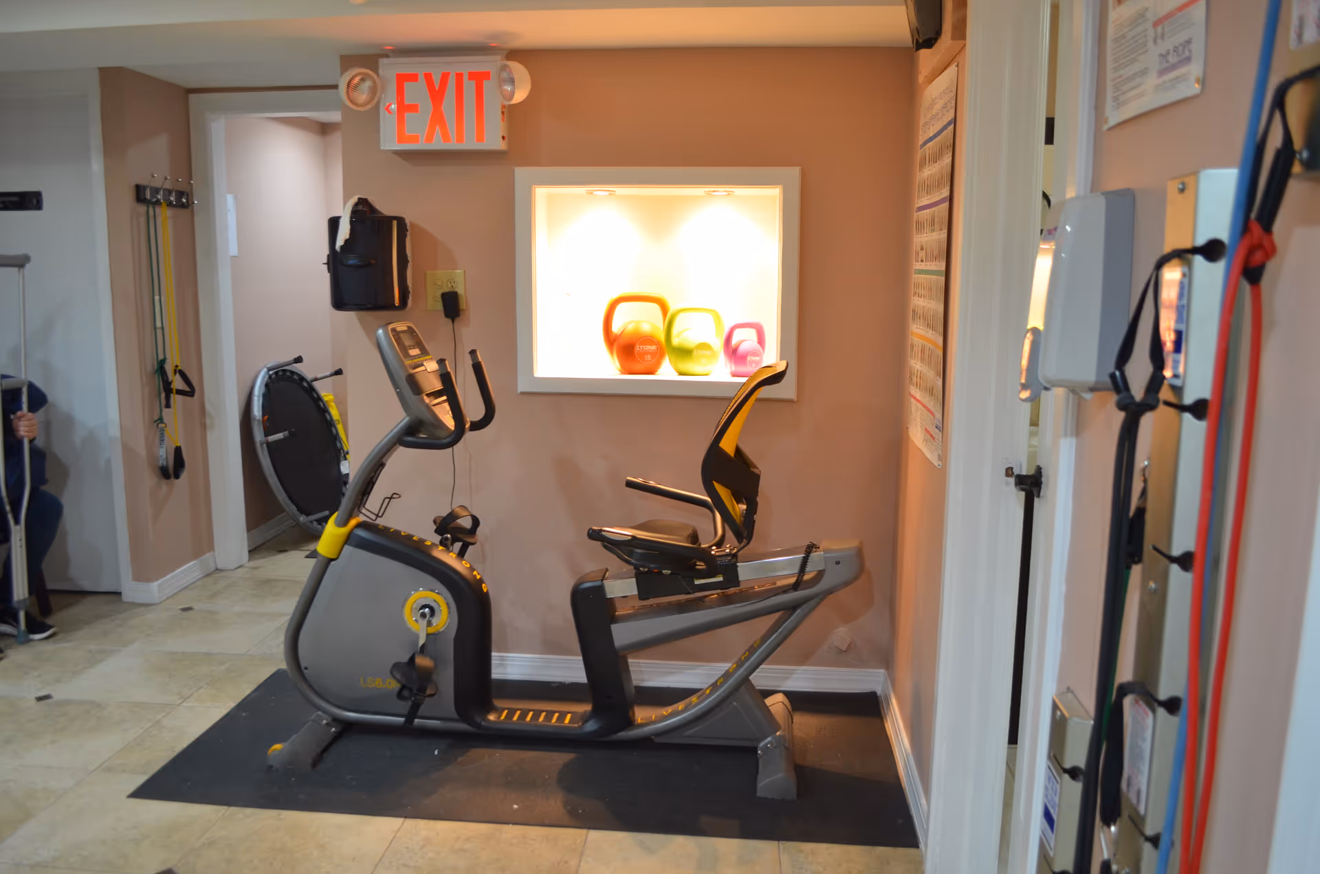 Recumbent exercise bike in a small rehab room with colorful kettlebells.