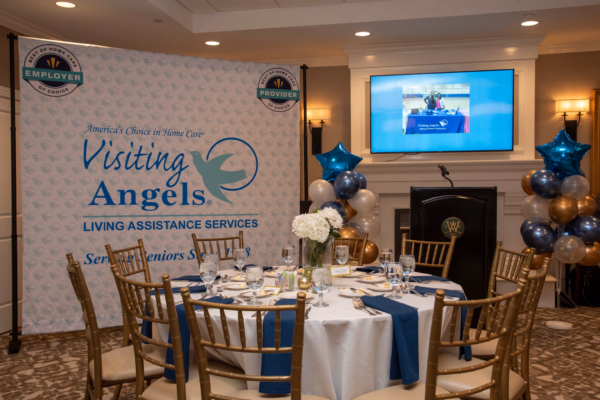 Indoor dining setup with a round table, chairs, banners, and balloons for Visiting Angels event.