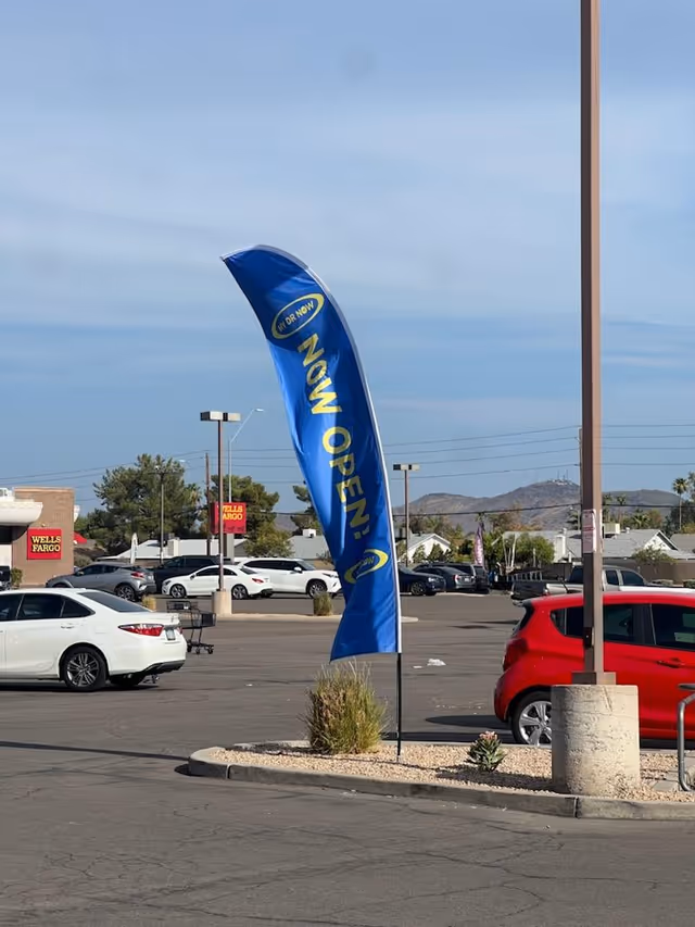 Blue banner reads Now Open in a parking lot with several parked cars.
