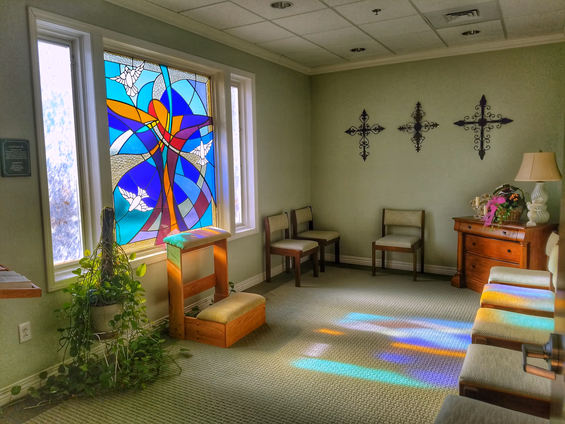 Sunlight streams through a colorful stained-glass window onto a quiet, furnished interior room.