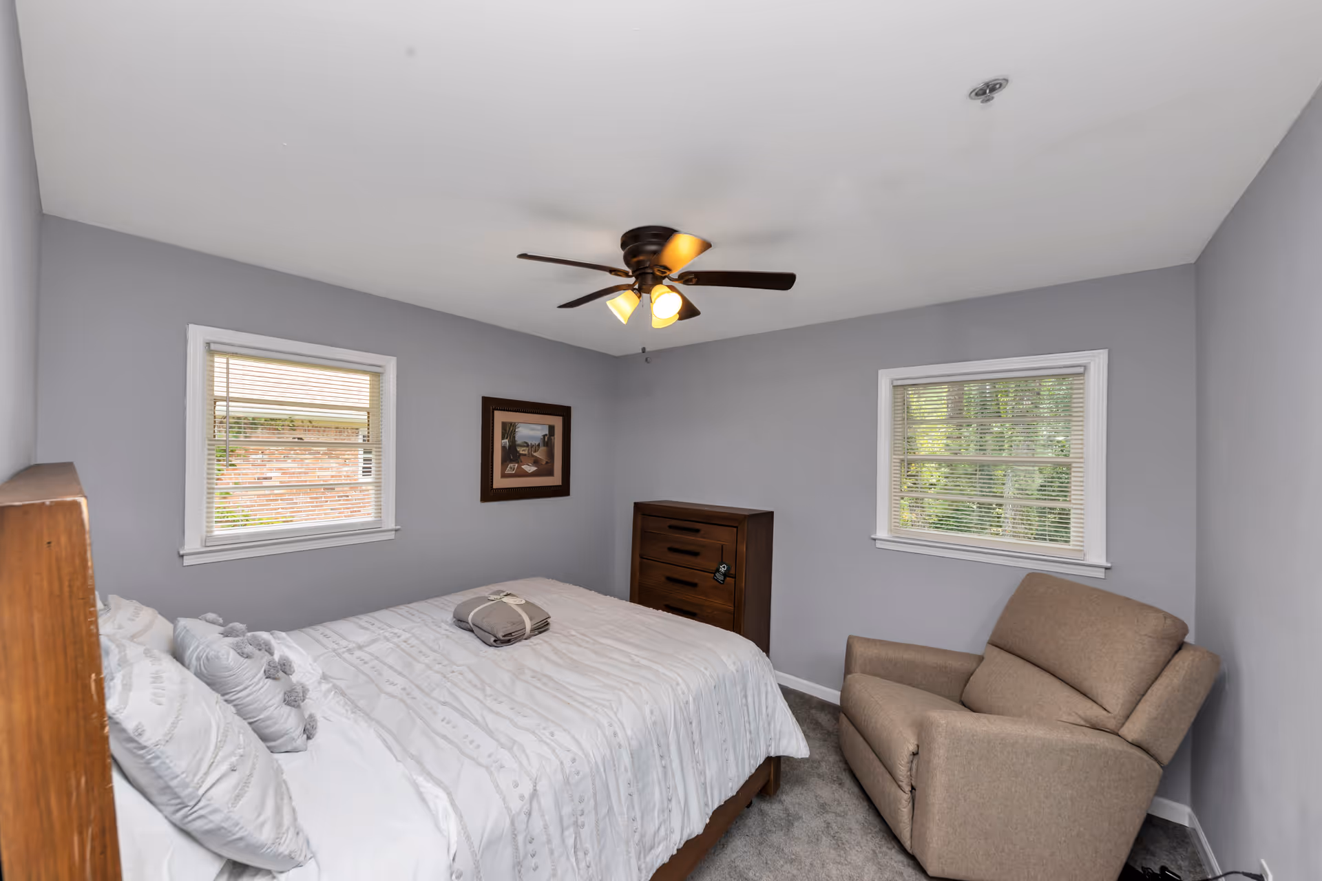 Bright, compact bedroom with bed, windows, dresser, and beige armchair in soft gray walls.