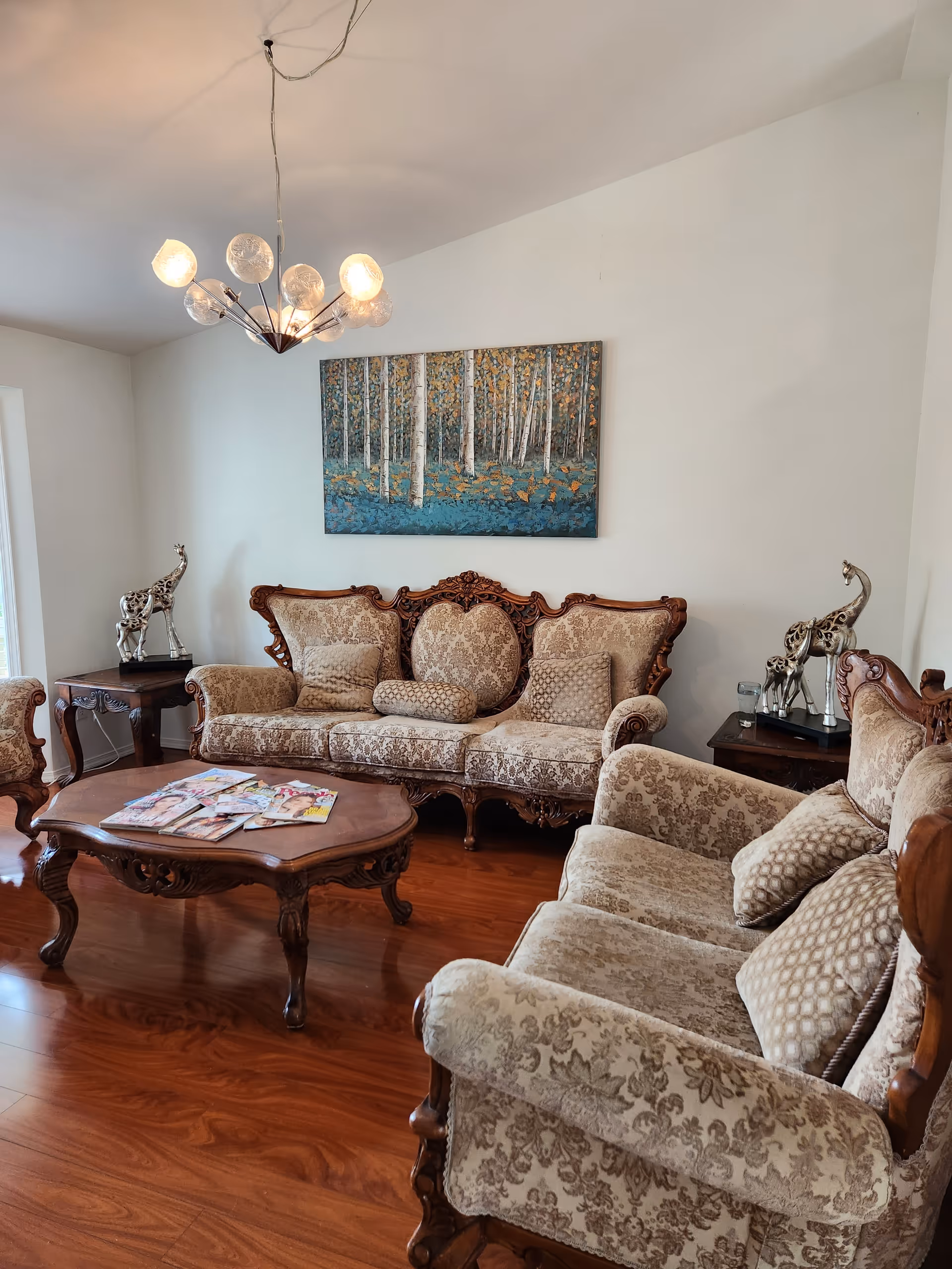 Elegant living room with ornate sofas, a wooden coffee table, chandelier, and forest painting.