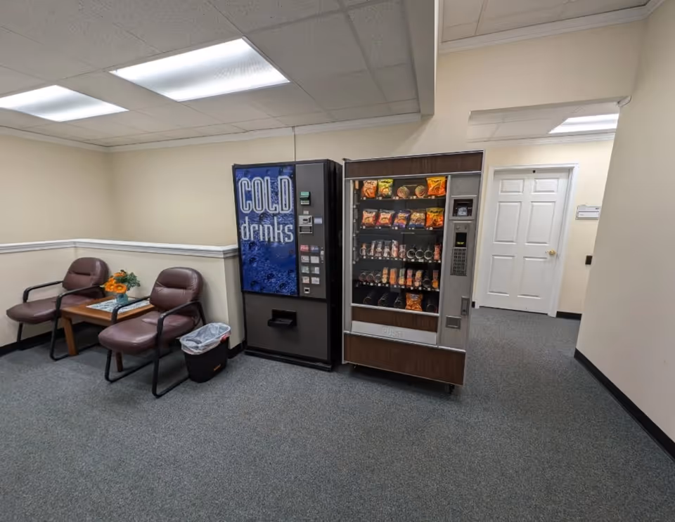 Two brown chairs with a small table sit beside vending machines in a carpeted hallway.