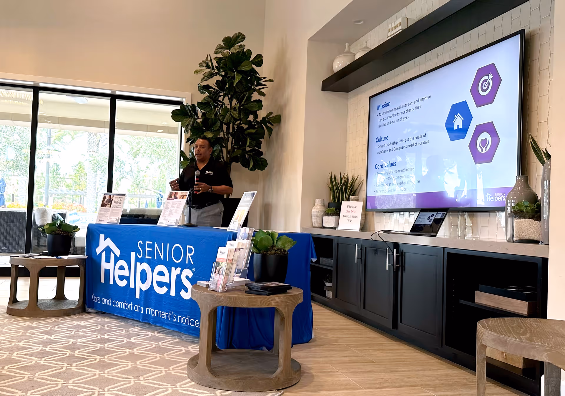 Presenter beside Senior Helpers banner in a modern reception area with a large screen.