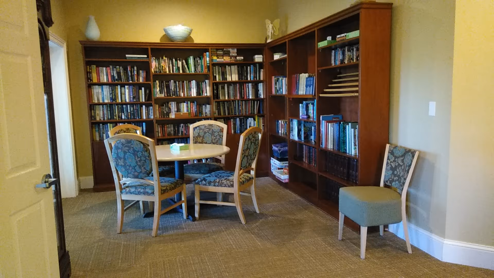 A cozy library in a senior living facility with a round table and blue upholstered chairs.