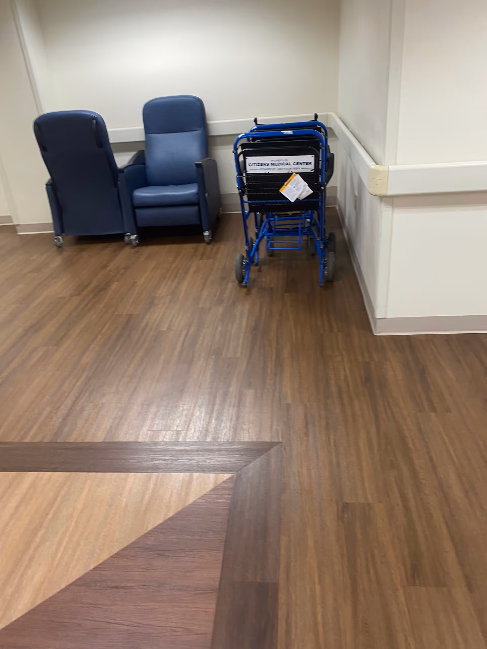 Blue chairs and a blue wheelchair cart occupy a corner in a hospital hallway