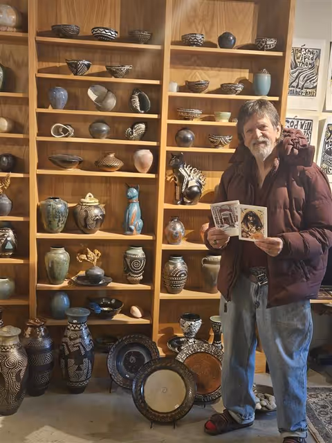 A man stands beside wooden shelves filled with pottery and holds magazines.