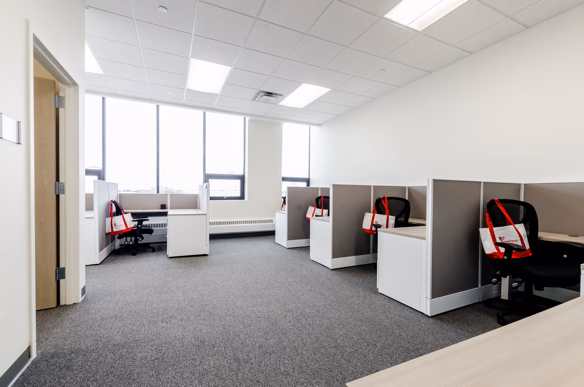 Bright office cubicles with desks, chairs, and red bags along a wall.