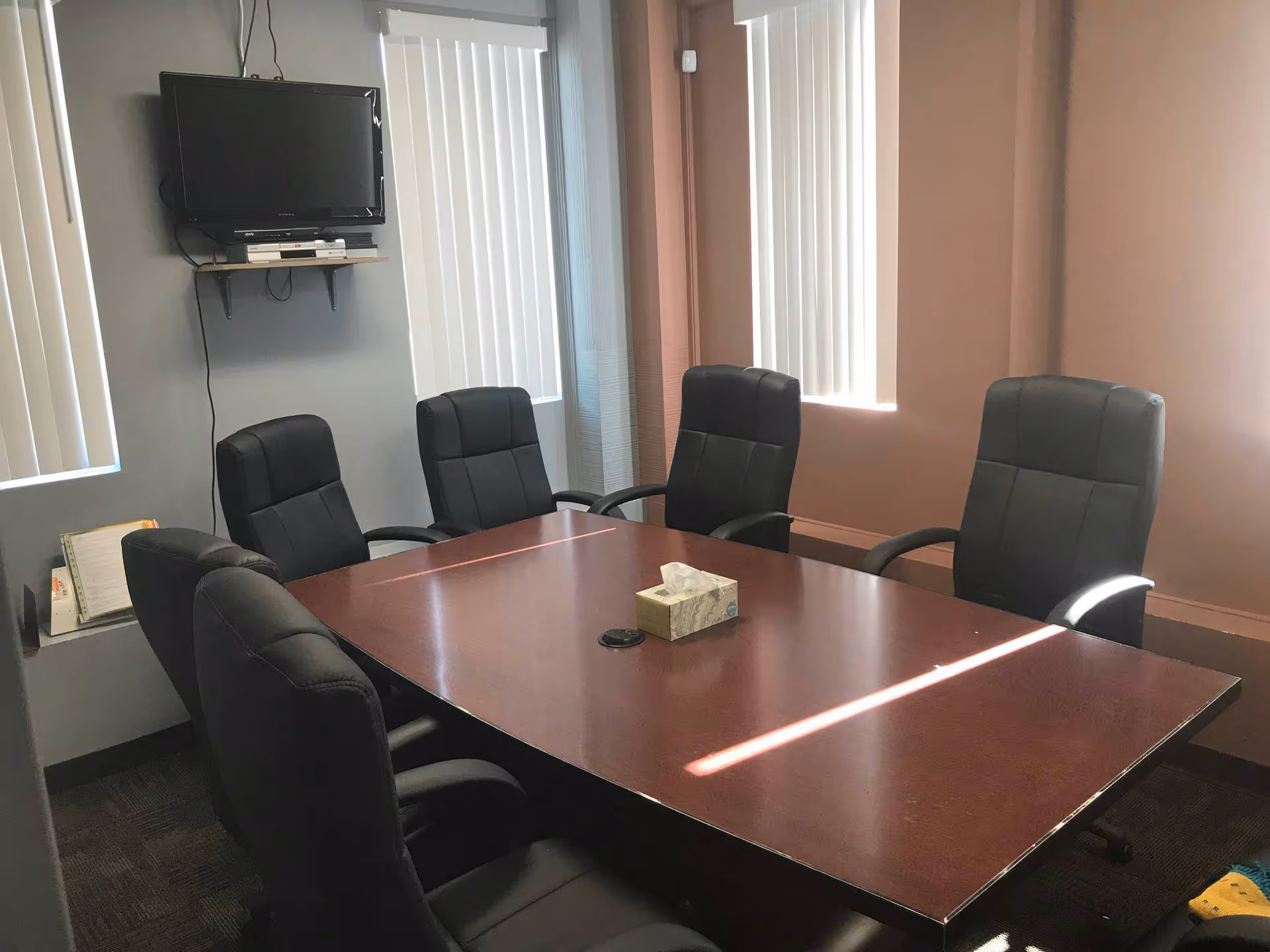 A conference room with dark wood table, black chairs, wall-mounted TV, and tissue box.