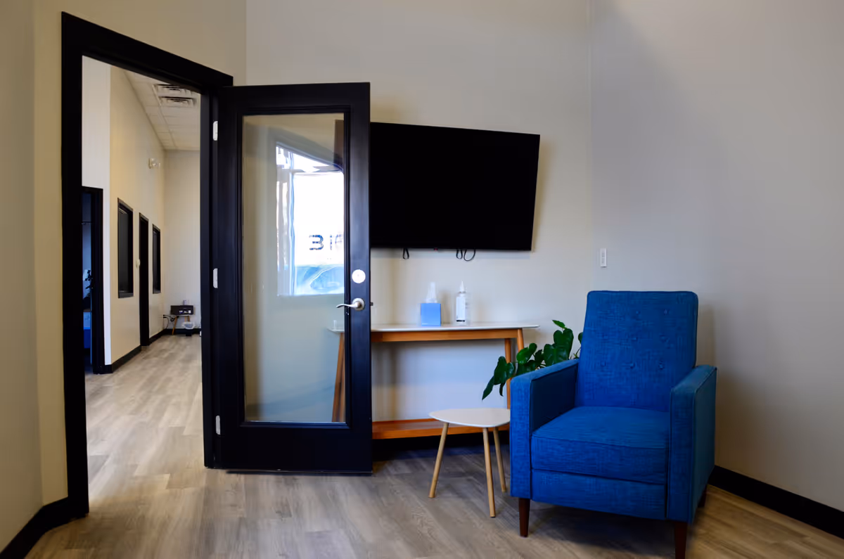 Glass door entry opens to hallway; TV, blue armchair, and small table in lounge.