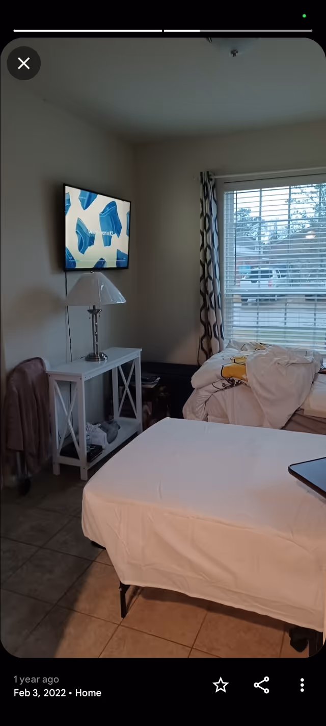 A bedroom with a bed, nightstand, wall-mounted TV, lamp, and large window.