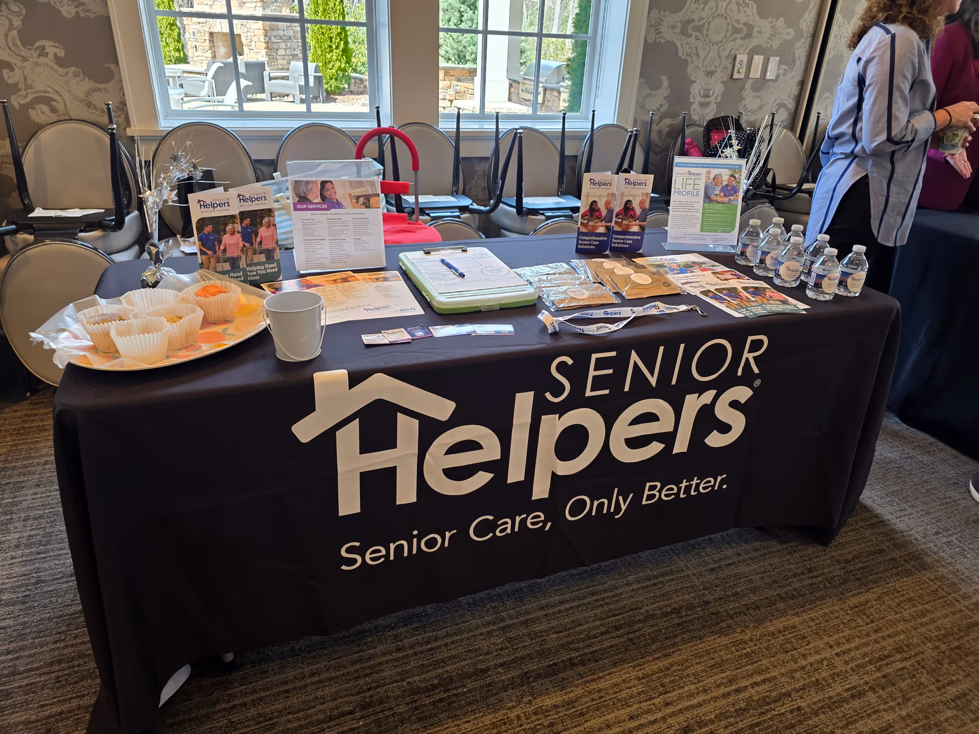 Table for Senior Helpers with brochures, cupcakes, water bottles, and chairs against a bright room.