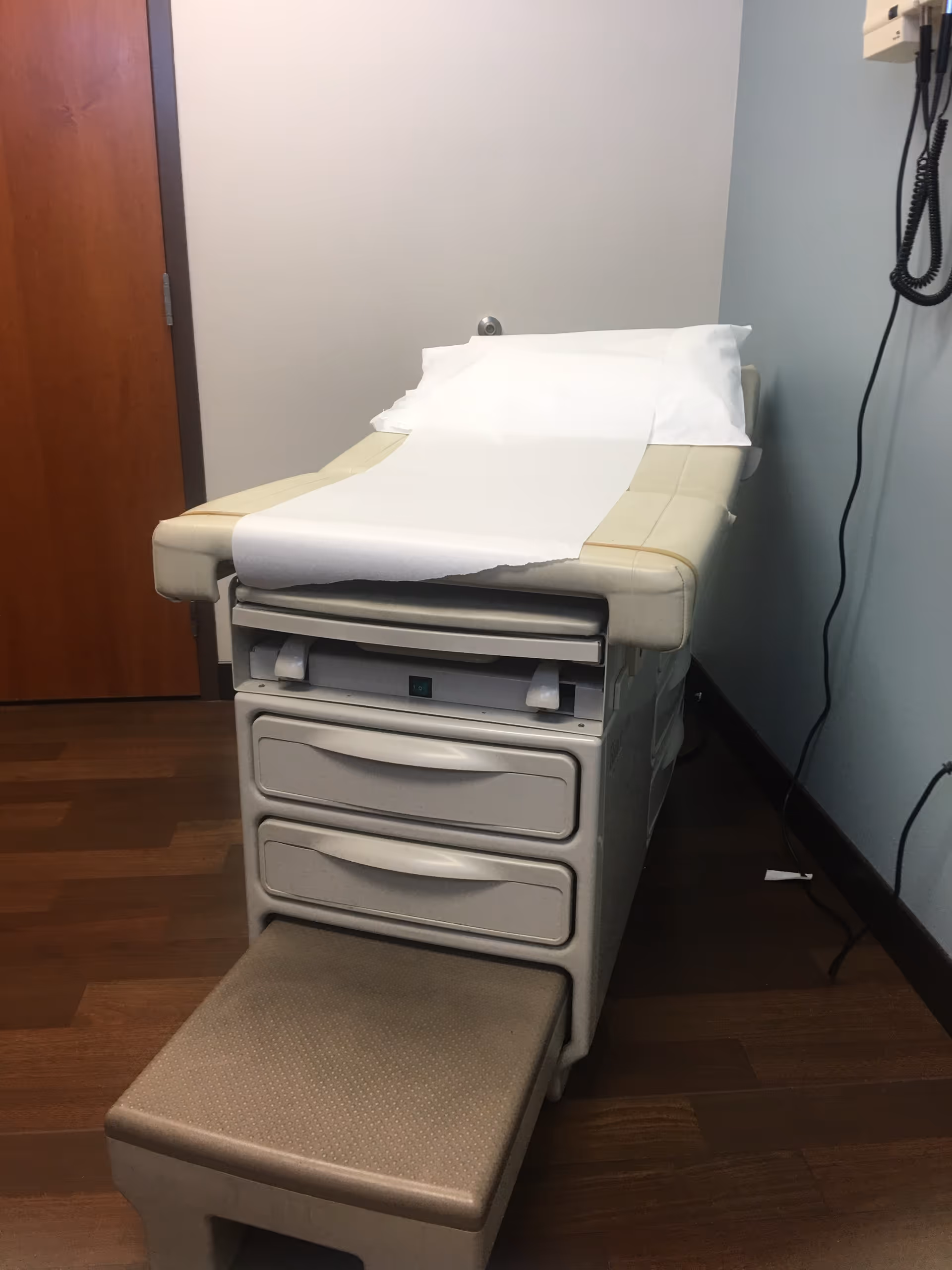 Examination chair with paper sheet and drawers in a small clinic examination room.