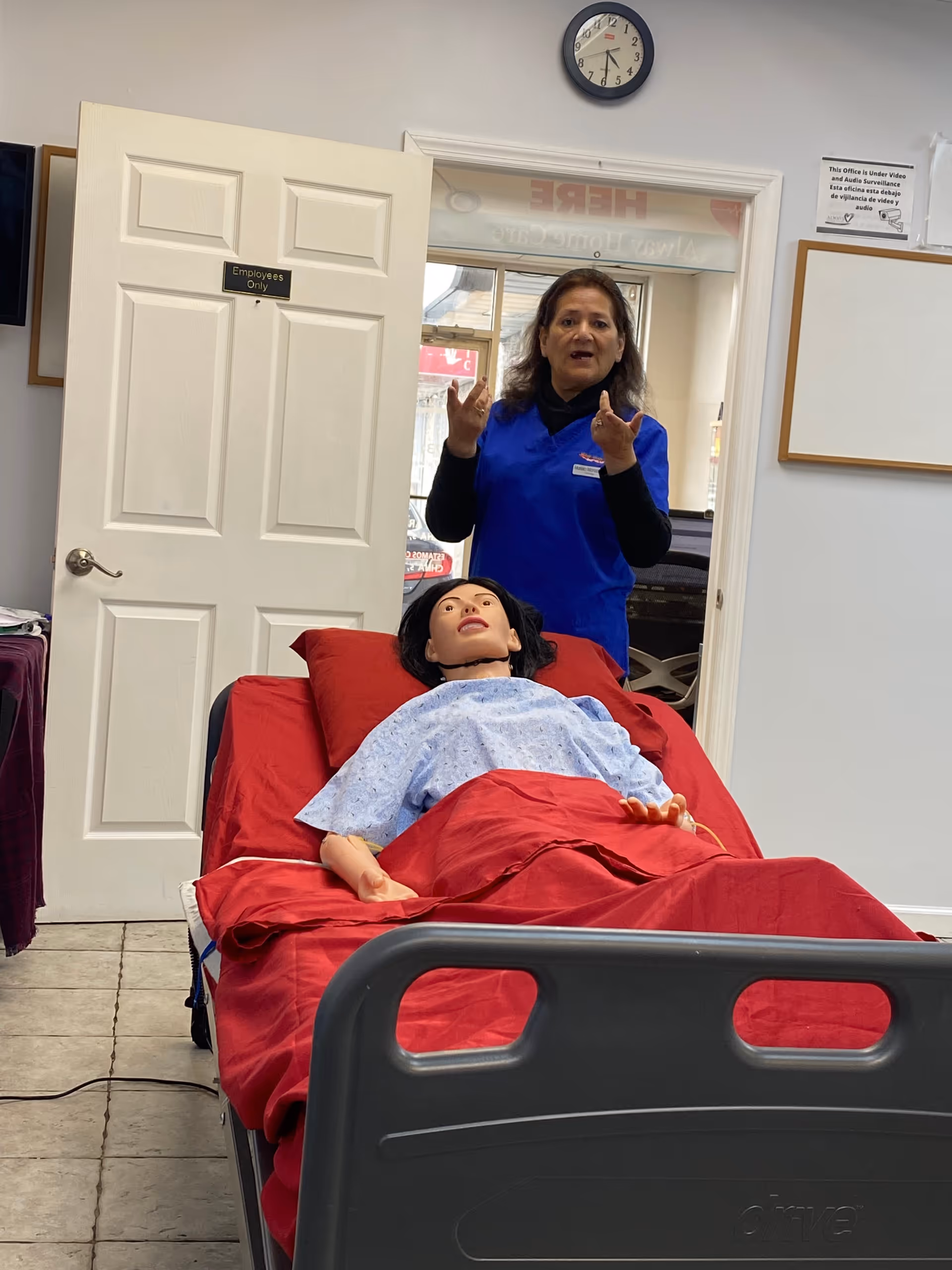Caregiver in blue scrubs explains to a medical mannequin on a hospital bed.