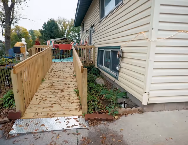 Wood ramp leads to a sunny outdoor deck with striped seating beside a beige house.