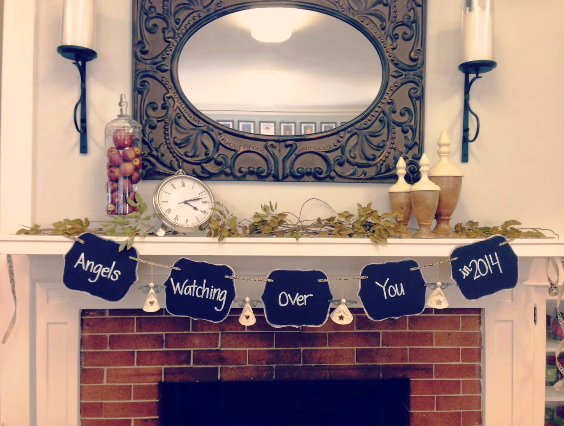 Ornate mirror above a mantel with a clock, garland, and banner reading Angels Watching Over You.
