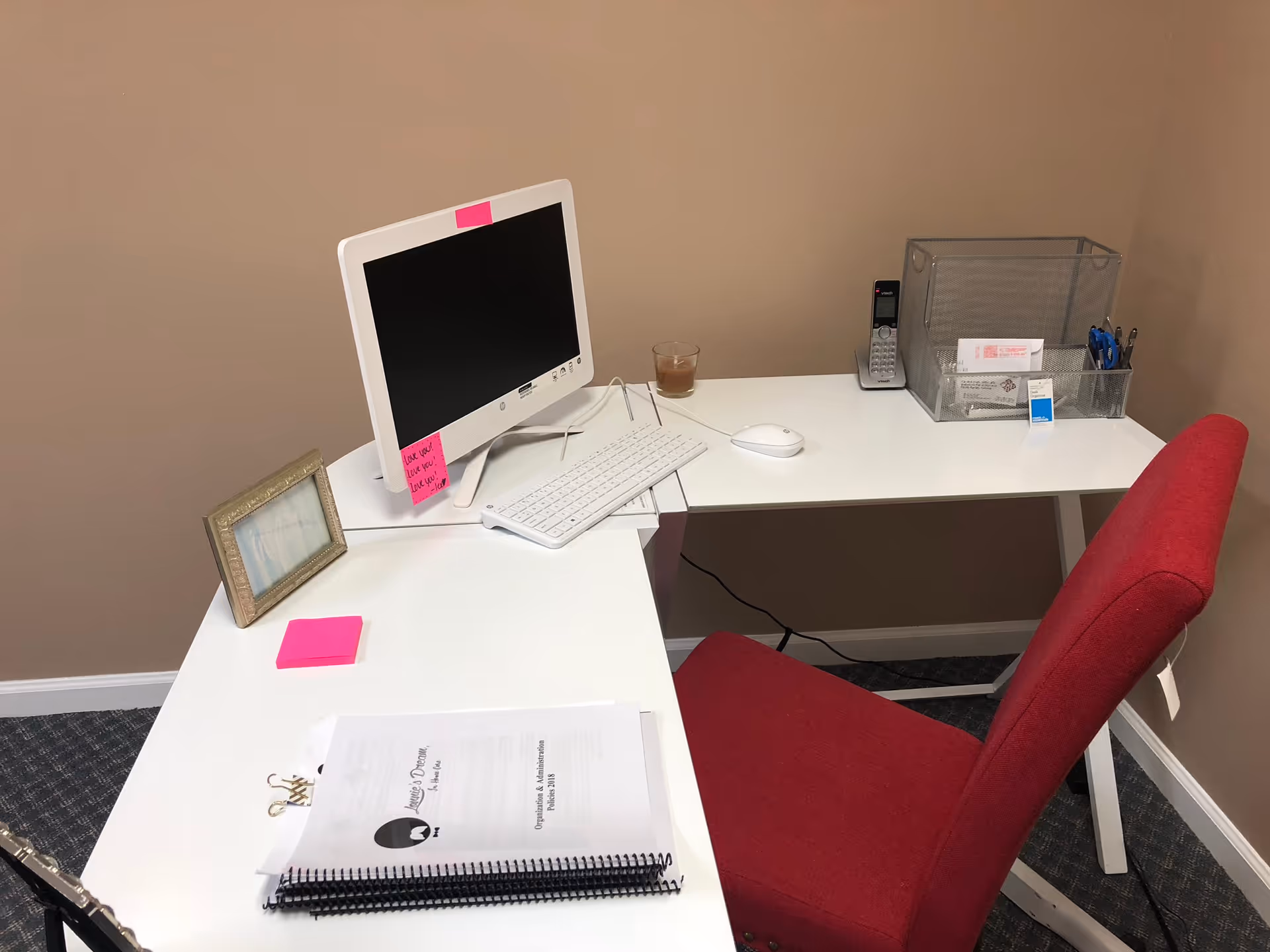 A white L-shaped desk with computer, keyboard, mouse, sticky notes, and red chair in beige room.