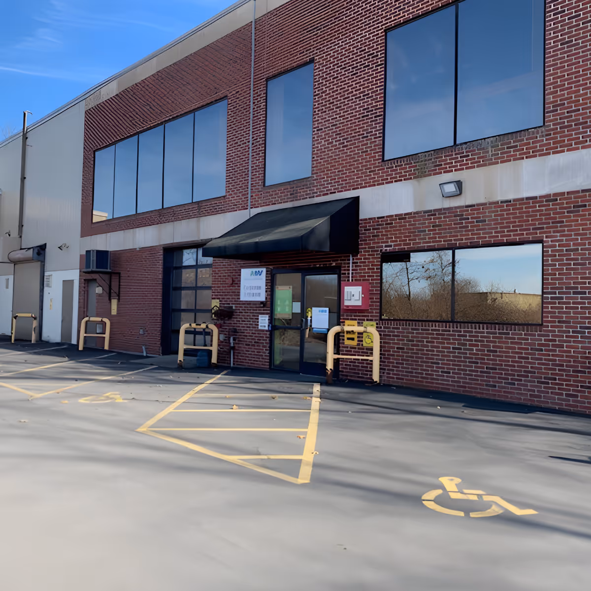Brick facility entrance with large windows, awning, and accessible parking in a sunny lot.