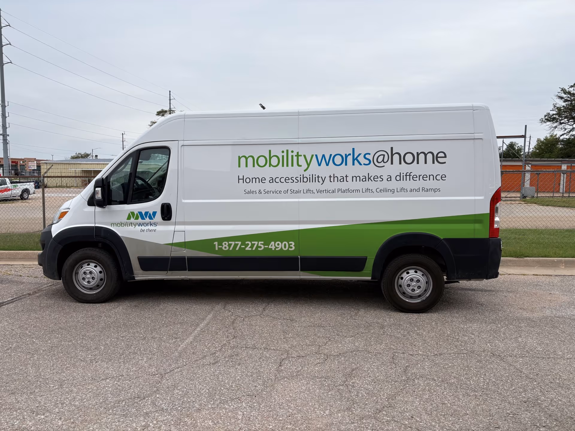 White MobilityWorks van with green stripe parked on a street beside a chain-link fence.