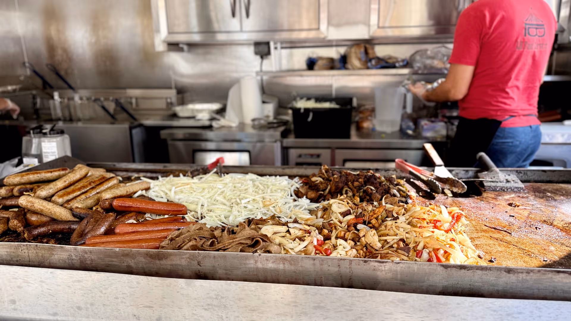 Commercial griddle with sausages, onions, peppers; caregiver in a red shirt cooks in a kitchen.