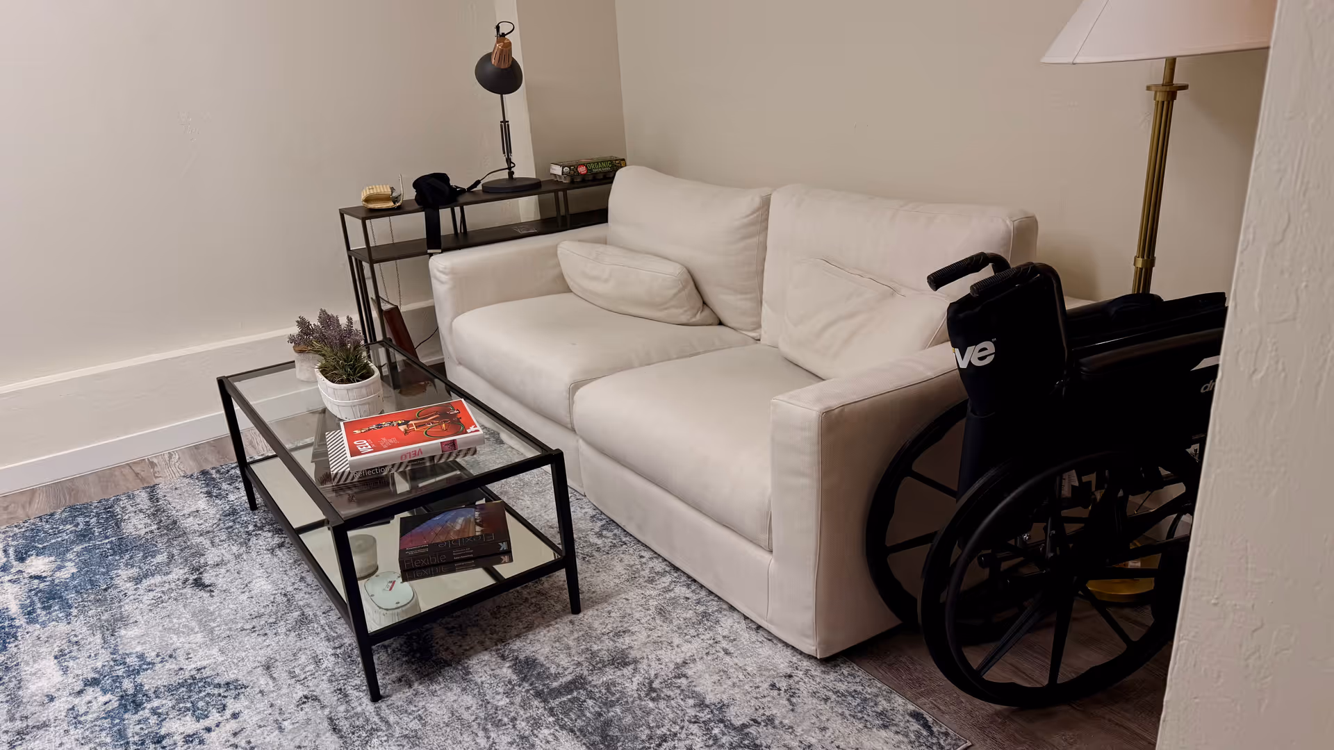 A small living room with a beige sofa, glass coffee table, rug, and wheelchair.